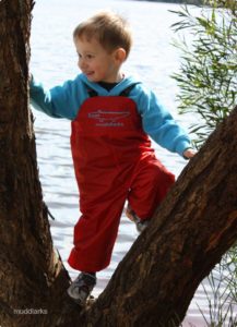 Boy climbing tree near a lake wearing red muddlarks® bib-n-brace overalls