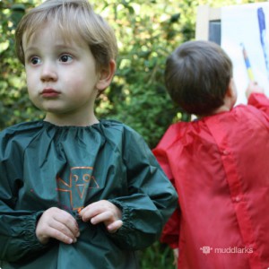 2 boys wearing muddlarks® childrens art smock painting outdoors, green smock shown from front, red smock shown from behind