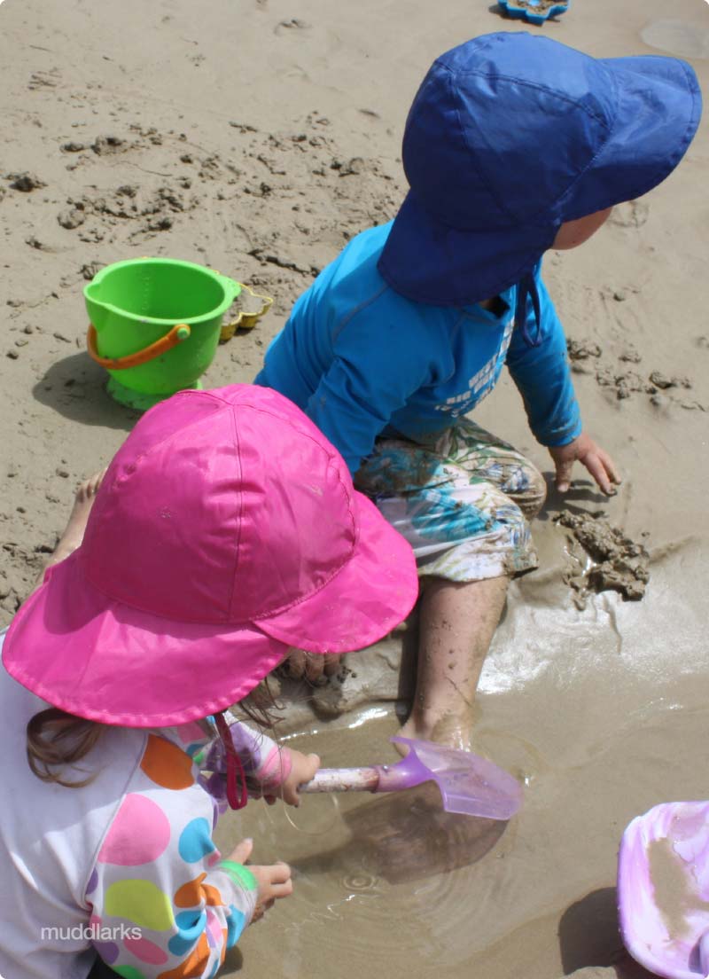 muddlarks® sou'wester hat kids playing in sand at the beach 2 kids playing in sand at the beach wearing muddlarks® sou'wester sun hat, one wearing pink hat and the other wearing blue